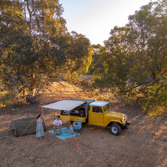 Yellow truck with a camper shell parked in a wooded area with people around.